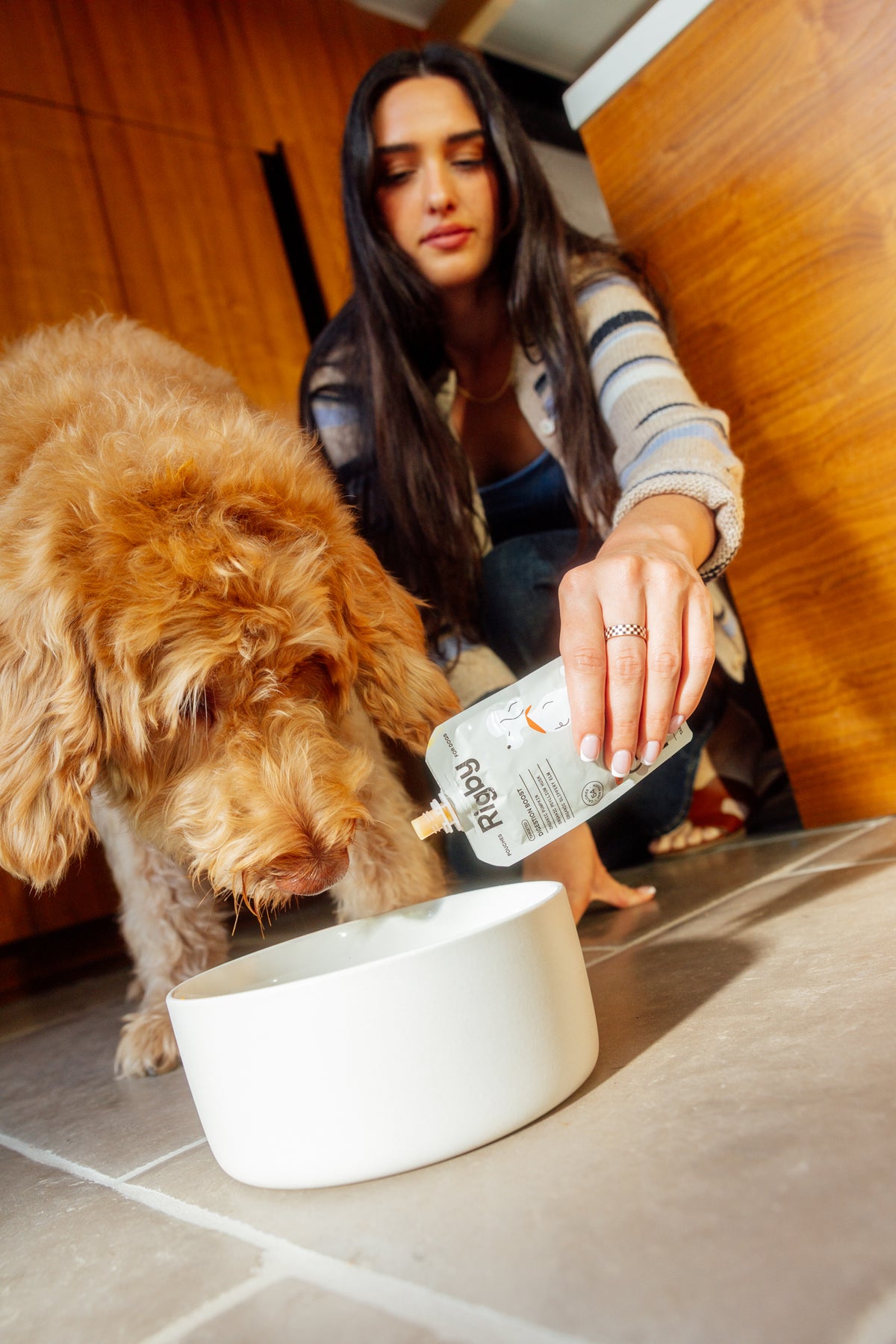 A woman in a striped sweater crouches on a tiled floor, squeezing a Rigby for Dogs pouch into a white bowl as a fluffy golden doodle leans in to eat.