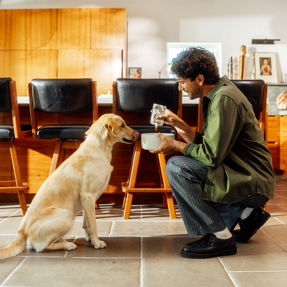 A curly-haired man in a green shirt crouches in a sunny kitchen, squeezing a Rigby for Dogs pouch into a white bowl as a yellow Labrador sits and licks its lips.