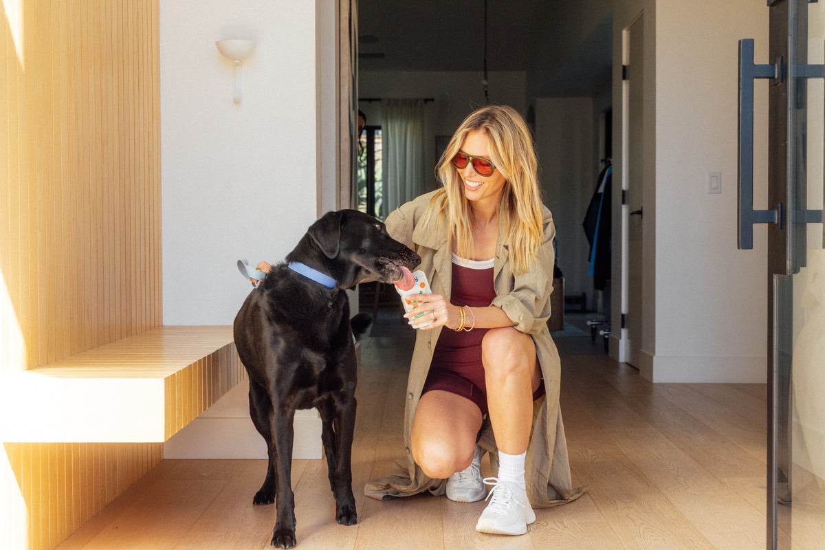 Woman sitting on a staircase with a black dog, both looking at the Rigby treat held by the woman.