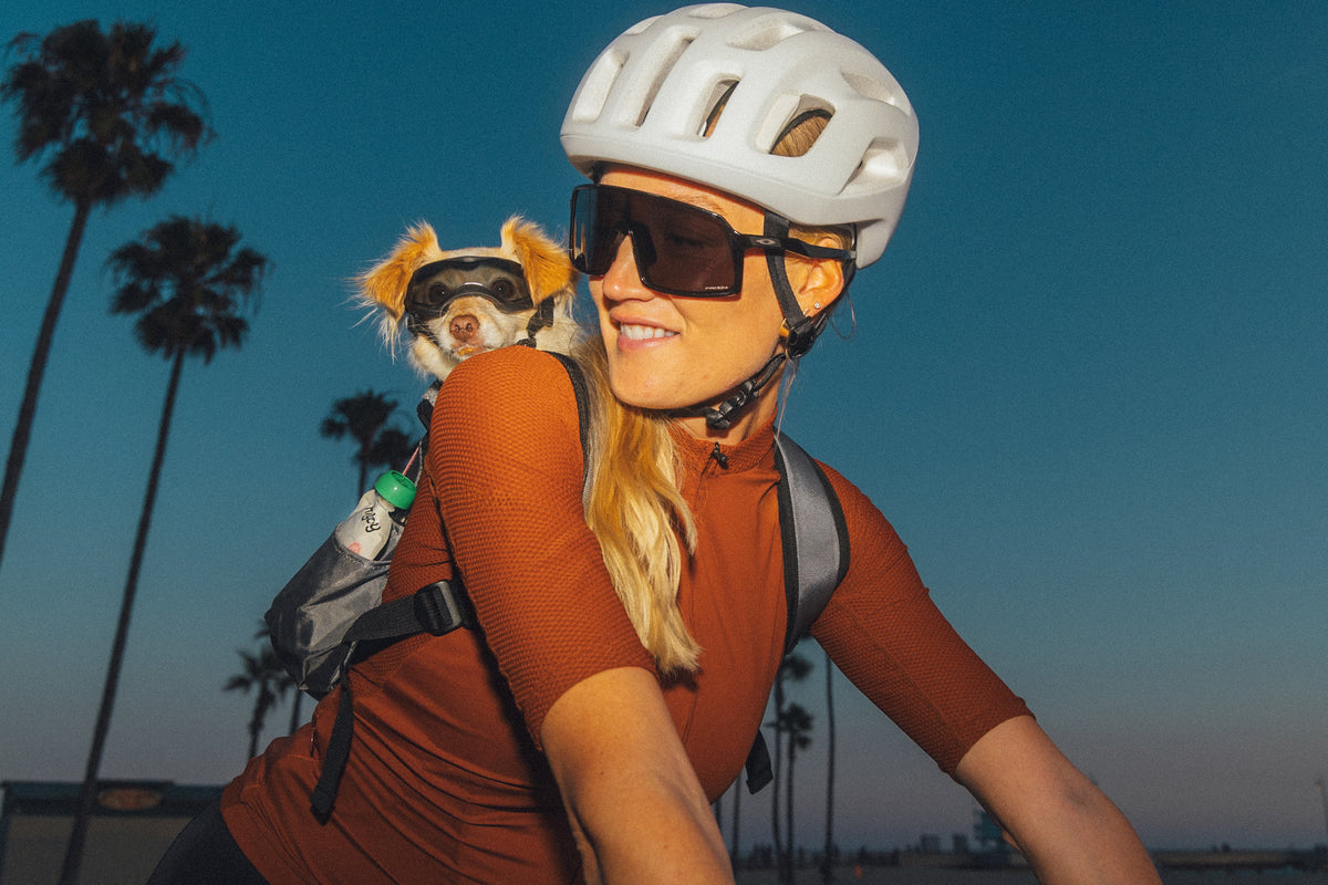 A smiling woman in a white bike helmet and sunglasses rides at dusk with a small dog in goggles peeking from her backpack, a Rigby for Dogs pouch tucked in the side pocket.