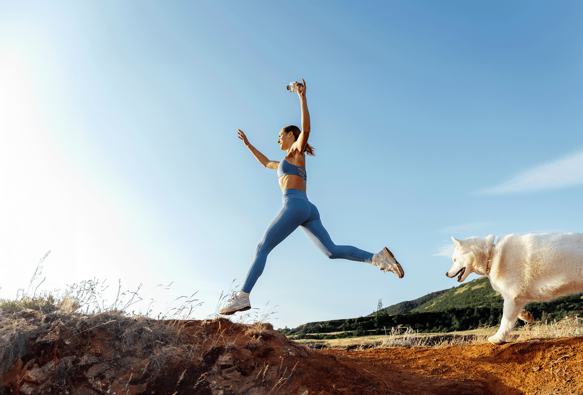 A woman in blue athletic wear leaping mid-air while holding a Rigby pouch, running alongside a white dog on a sunny dirt trail.